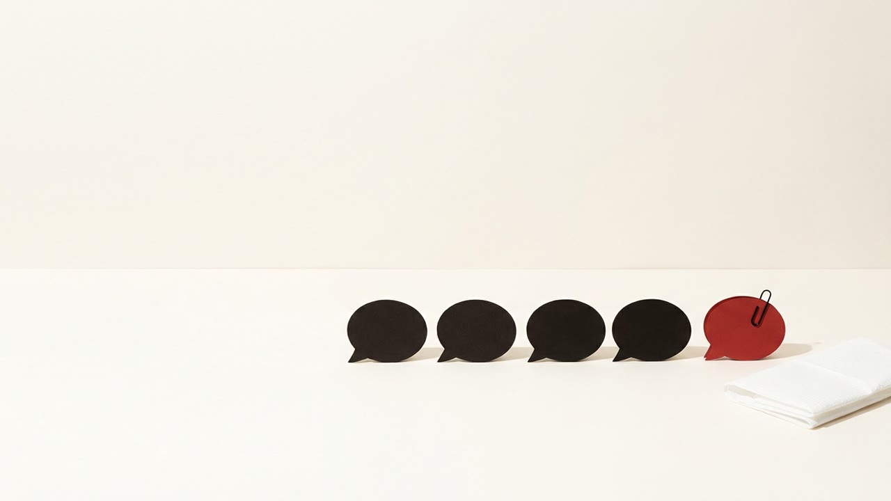 A row of black paper speech bubbles on a cream surface, the last one red and clipped shut with a paper clip, beside a folded white dental bib
