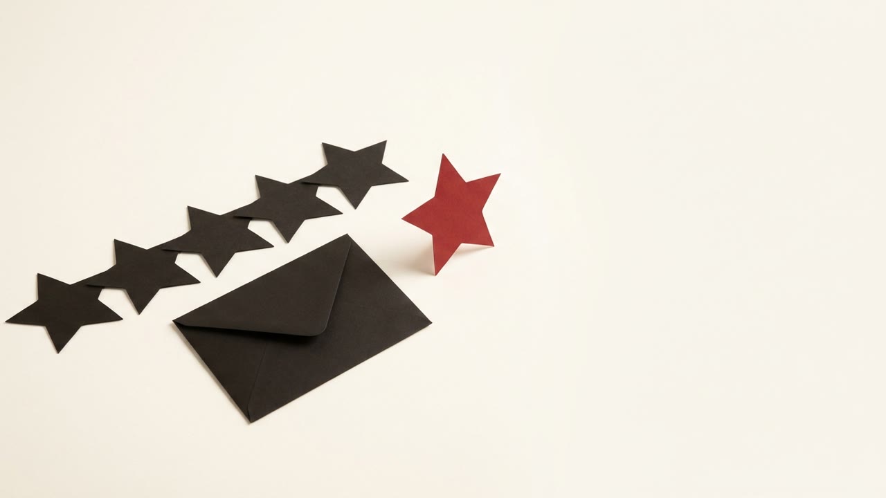 A diagonal row of black paper stars on a cream surface, with a red paper star standing upright and set apart from them, and a plain black envelope lying nearby