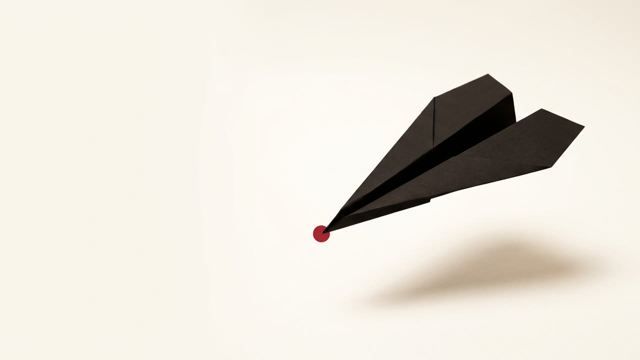 A black paper airplane folded from a single sheet, mid-flight over a cream surface, with one small red dot marking its nose