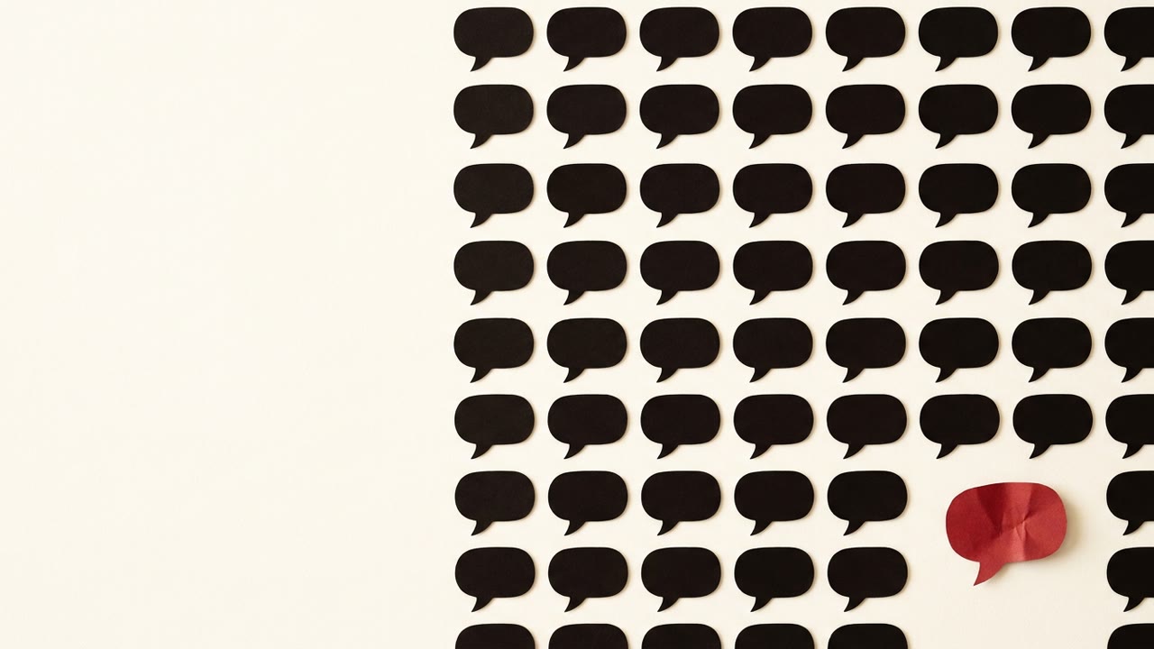 An overhead grid of identical black paper speech bubbles, with one folded differently and colored red, set apart from the uniform rows