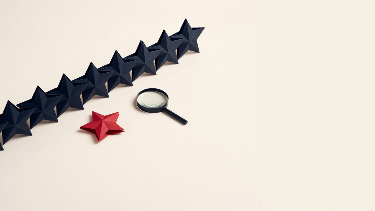 A cream surface with a row of black paper stars and one red star set apart, a black magnifier resting beside the row